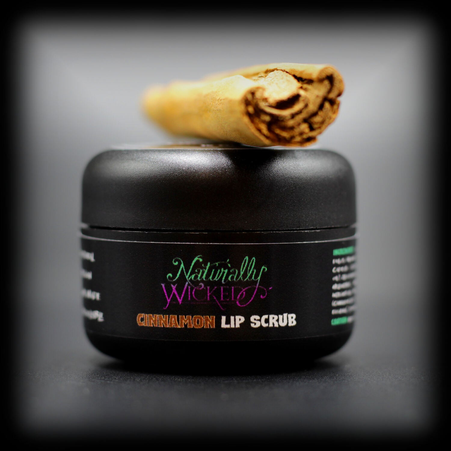 Naturally Wicked Cinnamon Lip Scrub In Luxury Black Container Beside a Fresh Spicy Cinnamon Stick