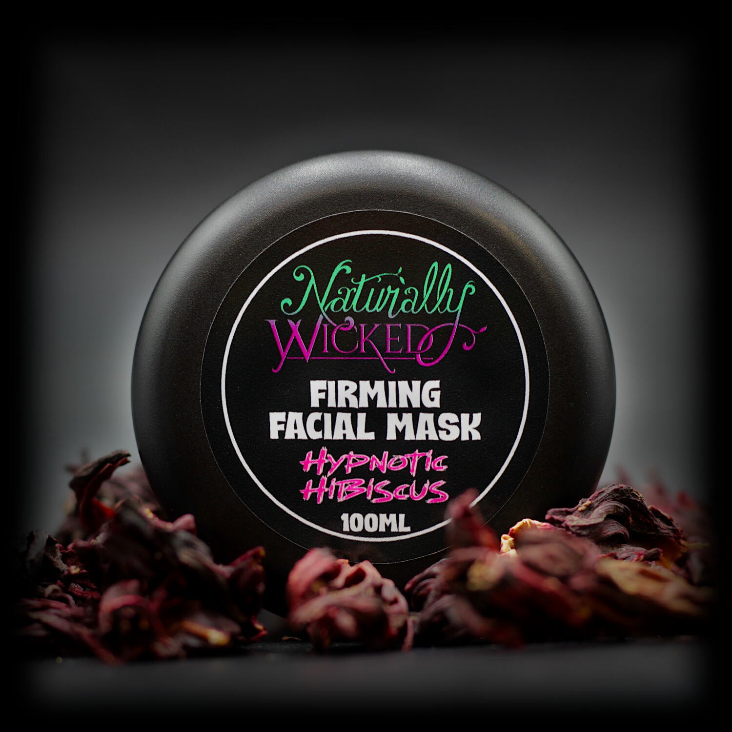 Naturally Wicked Hypnotic Hibiscus Firming Facial Mask in black tub, surrounded by dried hibiscus petals.