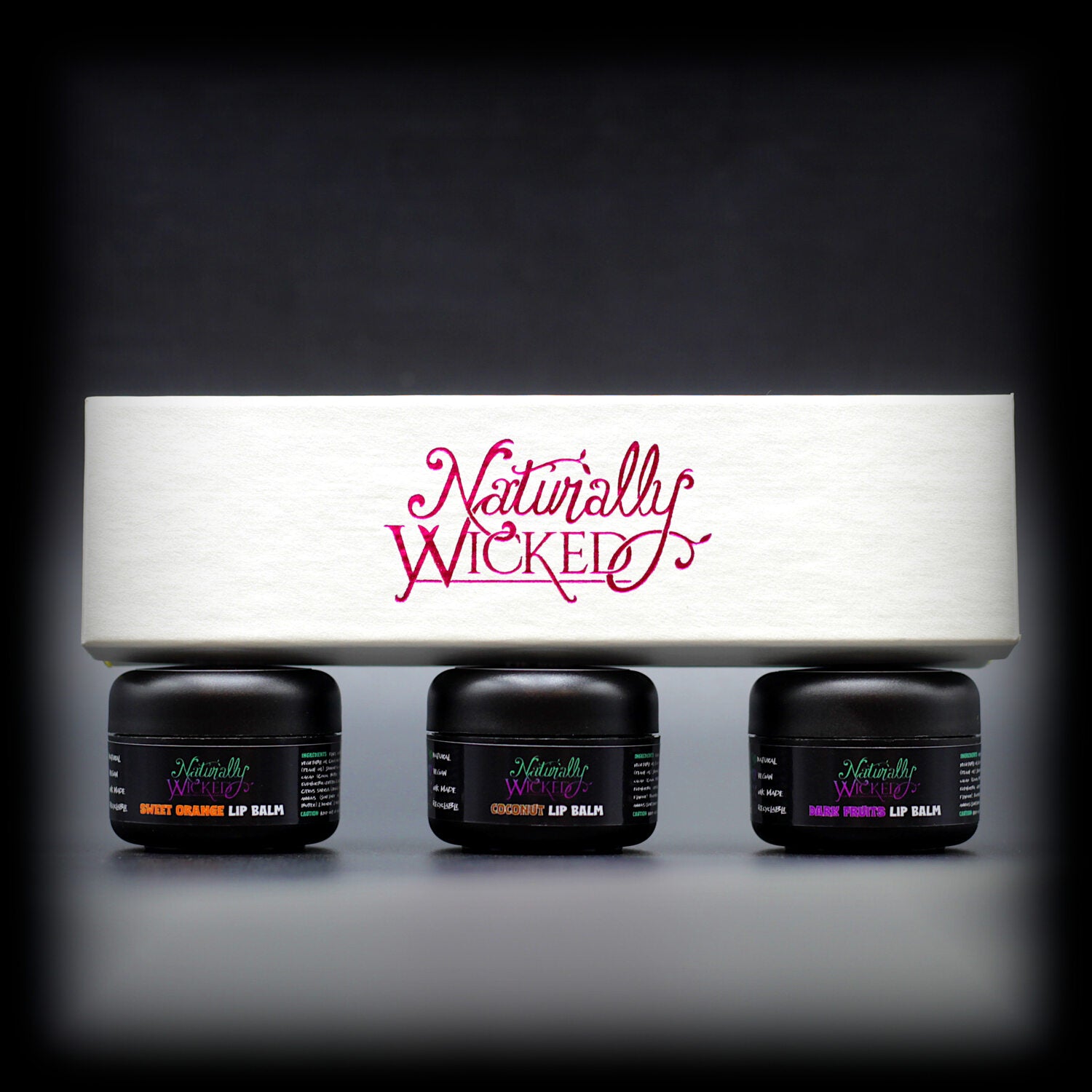 Naturally Wicked Lip Balm Trio Box On Top Of 3 Luxury Fruit Filled White Lip Balm Containers; Coconut, Dark Fruits & Sweet Orange